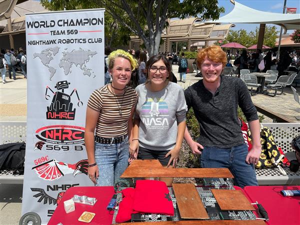 Three robotic students standing behind a table with a robot that folds a shirt